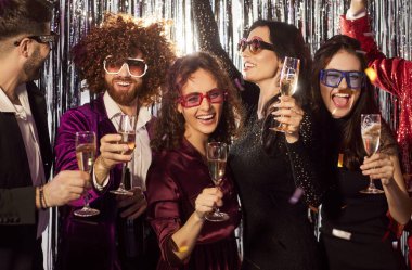 Partide müzik eşliğinde şampanya bardaklarıyla dans eden mutlu ve çeşitli arkadaşların grup portresi. Eğlence, neşe ve birliktelik, neşeli ve şenlikli bir atmosferi vurguluyor..