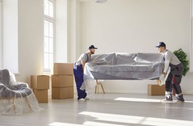 Oturma odasında ağır kanepe taşıyan iki genç servis çalışanı. Teslimat servisinden gelen erkek işçiler yeni eve mobilya yerleştiriyorlar. Taşınma günü ve taşınma konsepti.