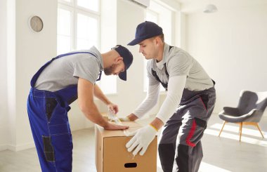 Genç erkek işçiler yeni bir daireye taşınmaya hazırlanan yapışkan bantla karton kutu paketliyorlar. Nakliye ekibi iş tulumuyla çalışıyor. Taşıma ve nakliye konsepti.