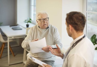 Yaşlı, beyaz saçlı bir adam muayenesi sırasında klinikte bir erkek doktorla konuşuyor. Yaşlı hastaların şikayetlerini dinleyen bir erkek doktor. Sağlık ve Tıp Konsepti.