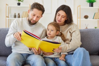 Evdeki kanepede oturan küçük tatlı kızlarıyla kitap okuyan mutlu bir aile. Gülümseyen ebeveynler sevimli çocuklarıyla vakit geçirip gülümsüyorlar. Birlikte boş zaman konsepti.