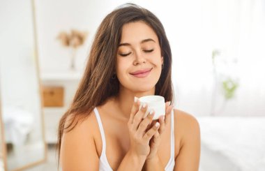 Neşeli genç bayan sabah cilt bakımının keyfini çıkarıyor. Gözleri kapalı, yüzünde nemlendirici yüz kremiyle gülümseyen beyaz kız cildini yumuşak, ışıl ışıl ve nemli tutuyor. Cilt bakımı.