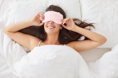 Sabahları rahat, modern bir yatak odasında beyaz çarşafların altında uyuyan, gülümseyen, maske takan genç bir kadının en güzel portresi. Evde dinlenen huzurlu bir kız. Yeni gün konsepti.