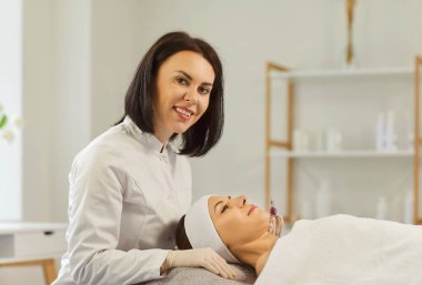 Kameraya bakan genç bir kozmetik uzmanı güzellik salonundaki kanepede uzanan güzel bayan müşteriye yüz bakımı prosedürü uygulayacak. Cilt bakımı ve kozmetoloji kavramı