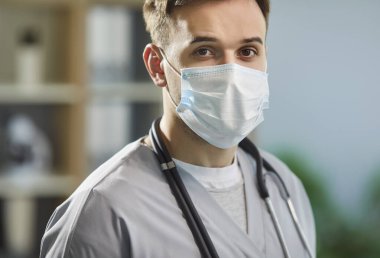 Yüz maskeli bir erkek doktorun yakın plan portresi. Gri önlük ve beyaz cerrahi maske takan genç bir adam tıbbi bir ofiste kameraya bakıyor.