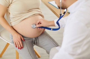 Klinikte ziyaret eden genç hamile bir kadını yakından inceleyen tıp jinekoloğu fetüsün kalp atış hızını steteskopla, hamile anne ve doğum öncesi bakımı ve sağlıklı hamileliği inceliyor.