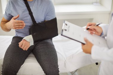 Kırık kollu bir erkek hastanın tıbbi muayenehane terapisi için doktoru ziyaret ederken çekilmiş portresi. Eli yaralı bir adam, koltukta travmatologla oturuyor..