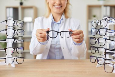 Genç, mutlu, gözlüklü kadın doktorun portresi. Ellerinde gözlük, klinikte çalışıyor. Çalışma masasında oturuyor. Oftalmoloji, vizyon ve göz sağlığı konsepti.