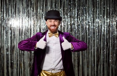 Mor kostümlü, gülümseyen mutlu bir adamın portresi. Akşam partisinde arka planda baş parmağını gösteriyor. Enerji ve neşe dolu bir ortamda, kutlamanın her anında..