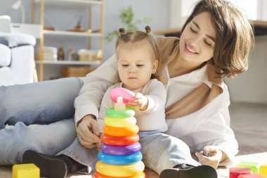 Pozitif anne ve genç kız evde yerde oturmuş oyuncaklarla oynuyor ve renkli bir kule inşa ediyorlar. Neşeli bir aile bağı, boş zaman ve ortak bir eğlence anı..