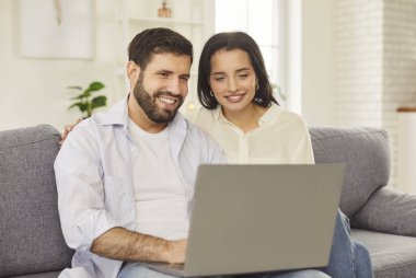 Genç, neşeli bir çift evdeki oturma odasında kanepede oturuyor. İnternet ve sosyal medya için dizüstü bilgisayar kullanıyorlar. Mutlu adam ve kadın kanepede dinleniyor. Haftasonu bilgisayarda video izliyorlar..