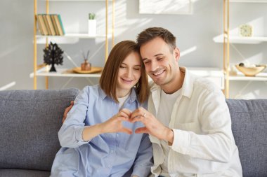 Oturma odasındaki kanepede birlikte oturan mutlu, genç çift elleriyle kalp şekli oluşturuyorlar. Memlekette sevgi, minnettarlık, şefkat ve güçlü duygusal bağları ifade eden neşeli erkek ve kadın..