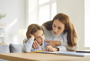 Neşeli anne babasının portresi. Evde masa başında oturup okul ödevi yapmaktan bezmiş küçük üzgün oğlunu destekliyor. Eğitim ve bilgi kavramı.