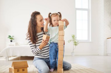 Mutlu anne ve çocuk birlikte vakit geçiriyorlar. Genç anne, küçük şirin kızına yardım ediyor. Ahşap bloklardan bir kule inşa ediyor. Yüzünde ciddi bir odaklanma ifadesi var.