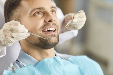 Modern diş hekimliği kliniğinde yatan genç ve mutlu bir erkek hastanın dişlerini diş aletleriyle muayene edip tedavi eden dişçiye yakın durun. Diş sağlığı, hijyen ve tıp konsepti.