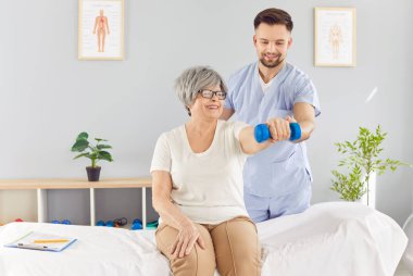 Doktor terapist, yaşlı bir hastaya rehabilitasyon ve fizyoterapi seansında yardımcı olur. Kol ve eklem egzersizlerine bir halterle odaklanır. Terapi gerilmeyi ve kas gücünü arttırır..