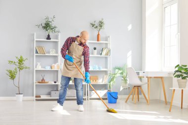 Neşeli adam evini temizlemekle meşgul, yerleri özenle süpürüyor. Ev işlerinin bir parçası olarak süpürge kullanıyor ya da temizlik yapıyor, ekipmanlarla temiz ve düzenli bir yaşam alanı sağlıyor..