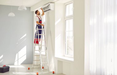 Modern klimayı içeride temizleyen profesyonel elektrikçinin tam boy portresi. Gülümseyen genç teknisyen yeni daireye klima tamir ediyor ya da kuruyor..