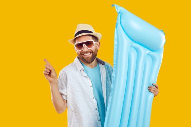 Sarı arka planda duran, elinde mavi su yatağı olan, yaz indirimi reklamını yapan ve parmağını havaya kaldıran yaz kıyafetleri içinde mutlu bir adam. Tatil, tatil, plaj, reklam konsepti