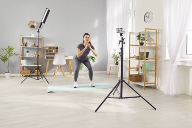 Fitness sportif vlogger, fit kadın spor etkileyicisi ev ilhamında çömelme videosu yapıyor, profesyonel koçluk danışmanlığı öneriyor, uzman ipuçlarını akıllı telefon kamerasıyla kaydediyor, motivasyon eğitimi alıyor.