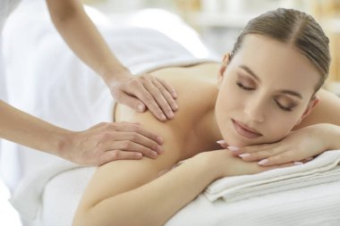 Masaj koltuğunda gözleri kapalı uzanan ve kaplıca salonunda omuz masajı yaptıran güzel bir genç kadın. Rahatlamış müşterilere stres, dolaşım ve sağIık terapisi sağlayan masaj terapisti