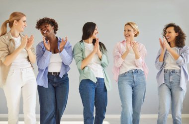 Moda kıyafetleri içinde beş farklı kadın, birlikte buluşuyor, gülümsüyor, el çırpıyor ve gri stüdyo arka planına karşı kapalı alanda birbirlerine bakıyorlar. Çok ırklı kadın modeller.