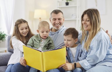 Beş çocuklu mutlu bir aile evde kanepede oturan çocuklarla kitap okuyor. Gülümseyen ebeveynler hafta sonunun tadını çıkaran çocuklarla vakit geçiriyorlar. İnsanlar ve aile eğlencesi konsepti.