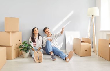 Mutlu, genç, gülümseyen bir çift yerde boş bir apartman dairesinde bir sürü karton kutuyla oturuyor. Yeni evin tasarım ve onarımını planlıyorlar. Taşınma günü, taşınma ve emlak konsepti.
