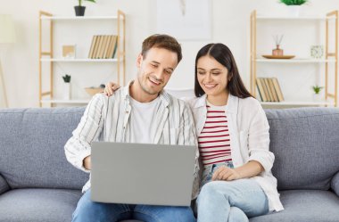 Oturma odasındaki rahat koltukta oturan mutlu, genç çift, kucaklaşıyorlar ve dizüstü bilgisayar kullanıyorlar. Neşeli aile hafta sonları online eğlencenin keyfini çıkarıyor, video seyrediyor ya da video görüşmesi yapıyor..