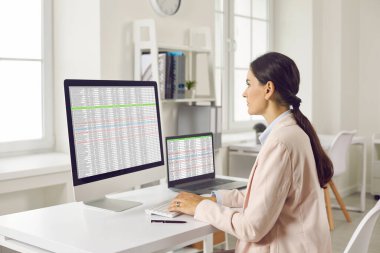 Masasında oturan, bilgisayarında masa ve çizelgeleri olan ve monitöre bakan konsantre genç bir iş kadınının yan görünüş portresi..