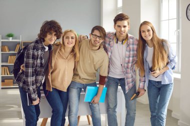 Bir grup mutlu gülen genç, sıradan kıyafetler içinde, dersten sonra modern sınıfta ayakta duruyorlar. Beş dost canlısı lise, üniversite ya da üniversite öğrencisinin birlikte kameraya bakan portresi.