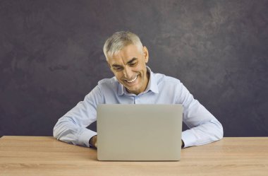 Orta yaşlı mutlu bir adam, masada oturup internette görüntülü konuşma yapıyor. Gülümseyen olgun iş adamı tipi dizüstü bilgisayarda çalışıyor iş yerindeki cihazlarda çalışıyor. İletişim, teknoloji kavramı.