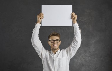 Siyah stüdyo arka planında gülümseyen bin yıllık beyaz bir adamın portresi boş bir fotokopi kağıdı içeriyor. Gözlüklü mutlu genç erkek pankart modeli satış anlaşması ya da tanıtım duyurusu yapıyor..