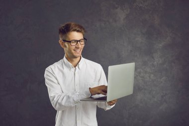 Güler yüzlü genç adam gri arka planda duruyor ve dizüstü bilgisayarına elinde tuttuğu metinleri yazıyor. Tişörtlü ve gözlüklü beyaz adam dizüstü bilgisayar ekranına rahatça bakıyor. İş konsepti.