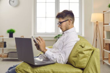 Evdeki kanepede oturan ve cep telefonu kullanan genç bir adam. Gözlüklü ciddi işadamının profil görüntüsü kanepeye dizüstü bilgisayarla oturmuş, cep telefonu ekranına bakıyor, numarayı çeviriyor ya da mesaj gönderiyor.