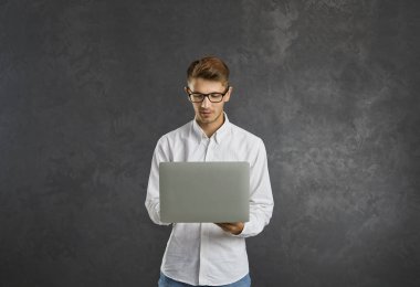 Başlangıç projesinde çalışan konsantre akıllı genç erkek programcı ya da çalışan. Elinde dizüstü bilgisayarla gri arka planda duran kendine güvenen beyaz bir adam. İş ve teknoloji kavramı.