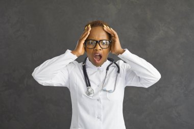 Stüdyo portresi şaşırmış beyaz önlüklü doktor ya da siyahi kadın hemşire gri arka planda izole edilmiş şok olmuş yüz ifadesine bakıyor.