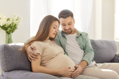 Beklenen ebeveynler hamile karnına dokunurken evdeki kanepede dinleniyorlar ve hamilelik yolculuğunun tadını çıkarıyorlar. Aşk, aile bağı ve yeni bebeğe kucak açmanın heyecanı..
