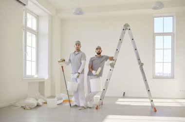 Boş bir odada merdivenin yanında duran, kovaları ve boya aletlerini tutan iki profesyonel ressam. Erkek işçiler kameraya bakıyor, yenilenmeye ve duvar boyamaya hazırlanıyor..