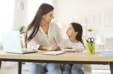 Mutlu anne-kız eğitimi, nişanlı öğrenci akıl hocası, öğretmen, bebek bakıcısı kız küçük öğrenciye masa başında yardım ediyor, öğreniyor, ev ödevi dersi, gülüyor, dizüstü bilgisayar kullanarak yazıyor.