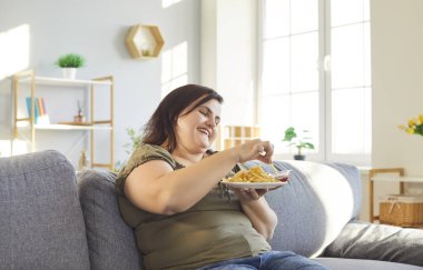Aşırı yağlı patates kızartması yiyen mutlu obez kadın koltukta oturuyor, sağlıksız fast food, lezzetli, tombul kilolu şişman şişman, iştahlı, aşırı kalorili kadın.