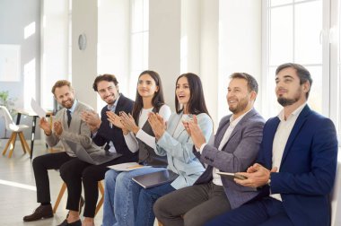 Resmi takım elbiseli bir grup iş adamı sunumlarını, çalışanlarını alkışlamak için ofiste oturuyorlar. Mutlu iş adamları el çırpıyor. Başarılı eğitim, takdir ve insan kaynakları ekibine teşekkür ediyorlar.