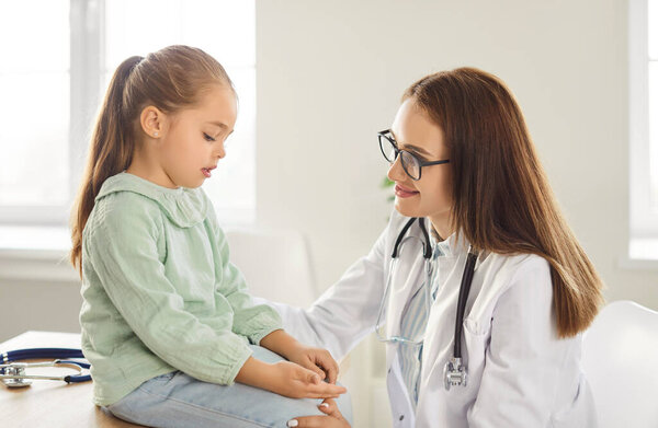 Smiling female doctor in glasses and white coat sitting in bright medical office and comforting little girl. Friendly pediatrician listening to kid patient complaints during checkup in medical clinic.