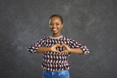 Gri stüdyo arka planında yalnız duran güzel kısa saçlı siyah kadın kalp hareketi yapıyor. Genç Afrikalı Amerikalı kadın elleriyle kalp şekli oluşturuyor, sevgiyi ve olumlu duyguları paylaşıyor.