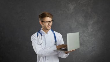 Tıp ve modern teknolojiler. Dost canlısı, modern ve zeki erkek sağlık hizmetleri uzmanı dizüstü bilgisayar kullanarak online hizmetler sunuyor. Sağlık önlüklü genç adam gri arka planda dikilirken daktilo çekiyor..