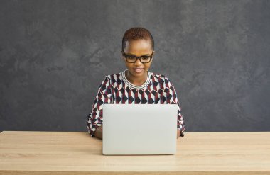 Çalışan, internetten alışveriş yapan ya da bilgisayarını kullanan arkadaşlarıyla mektuplaşan koyu tenli kadın portresi. Gri arka planda oturan, klavyeye yazı yazan odaklanmış genç bir kadın..