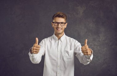 Mutlu bir adamın başarı, onay, yüksek kalite ya da iyi çalışma işareti olarak baş parmağını göstermesi. Gri arka planda dururken kameraya bakan gülümseyen beyaz adam. Pankart.