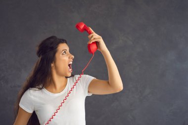 Sinirli bayan müvekkil telefonda tartışırken sinirlendi ve öfkeyle bağırdı. Gri arka planda duran beyaz bir kadın kırmızı sabit hatlı eski bir telefondan konuşuyor. Olumsuzluk kavramı.