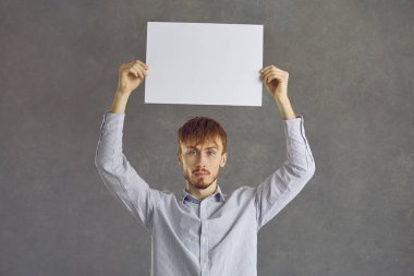 Ciddi bir adamın portresi, gri stüdyo arka planında dikilirken kafasında beyaz bir kağıt tutuyordu. Genç Kafkasyalı bilinçli adam oy veriyor ve konumunu bildiriyor. Boşluğu kopyala.