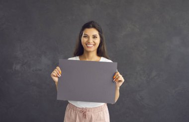 Gri arka planda duran boş gri bir kağıt parçası tutan güzel, mutlu bir kadın. Kafkasyalı tatmin olmuş bir kadının portresi fikrini ifade ediyor. Terfi konsepti. Düzen, kopyalama alanı.